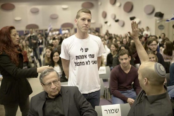 Young man challenges Naftali Bennett (foreground with raised hand). T-shirt reads "I have two moms and I don't apologize."